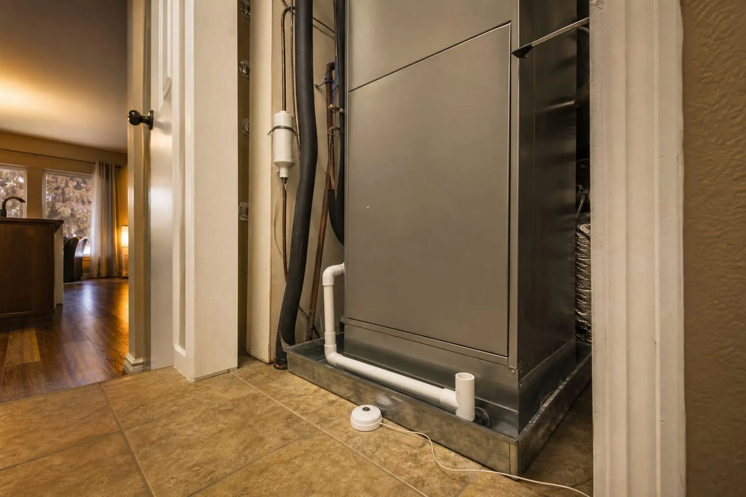 A white PVC condensate drain line and drain pan beneath an air handler in a residential utility closet, with a small water sensor puck on the floor beside it